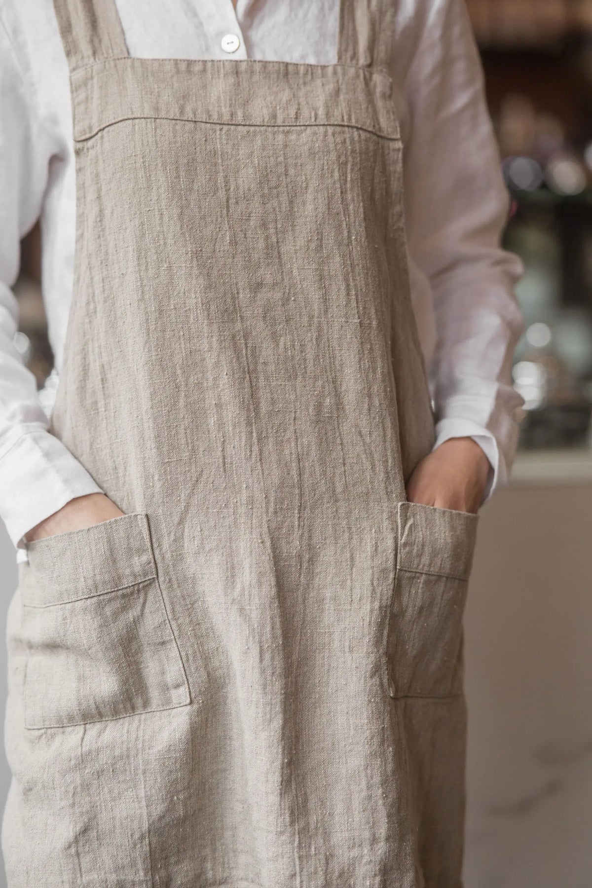 Close-up of two patch pockets on a natural linen apron, with hands resting inside each pocket, showing the linen's woven texture.