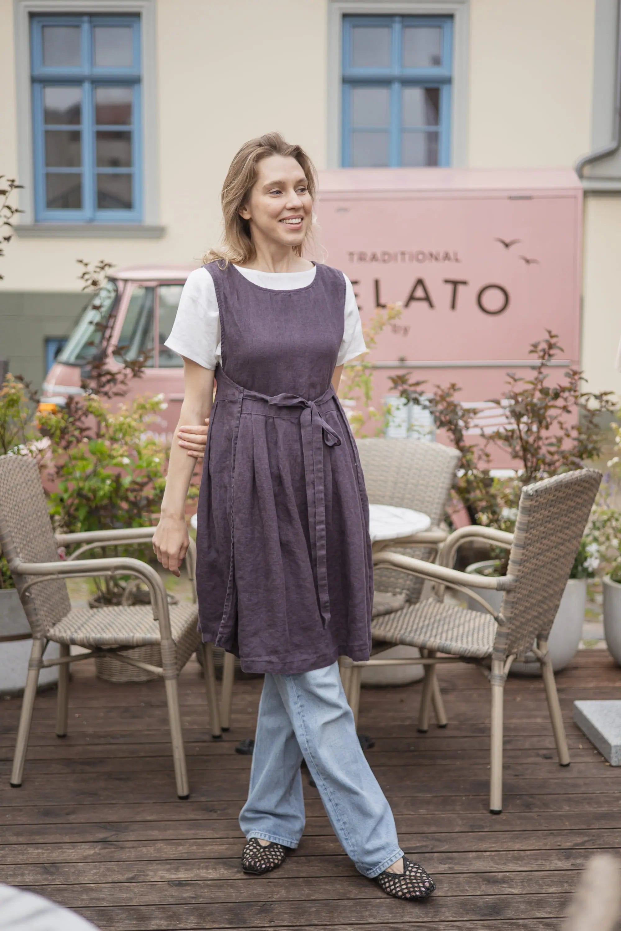 Front view of a woman laughing outdoors in a plum linen pinafore apron with a front tie bow, worn over a white t-shirt.