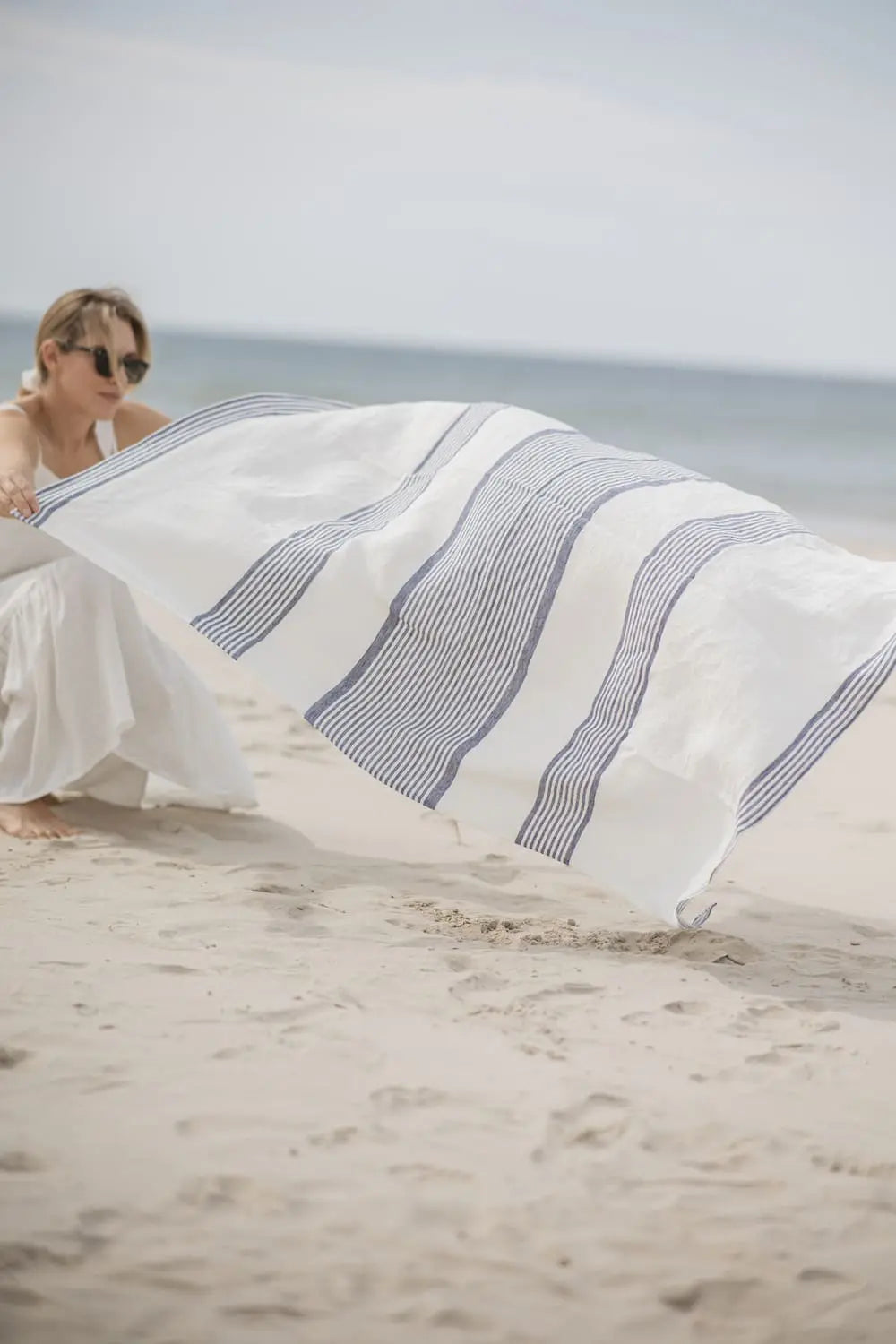 Woman unfolding a white and blue striped huckaback linen beach towel on the sand